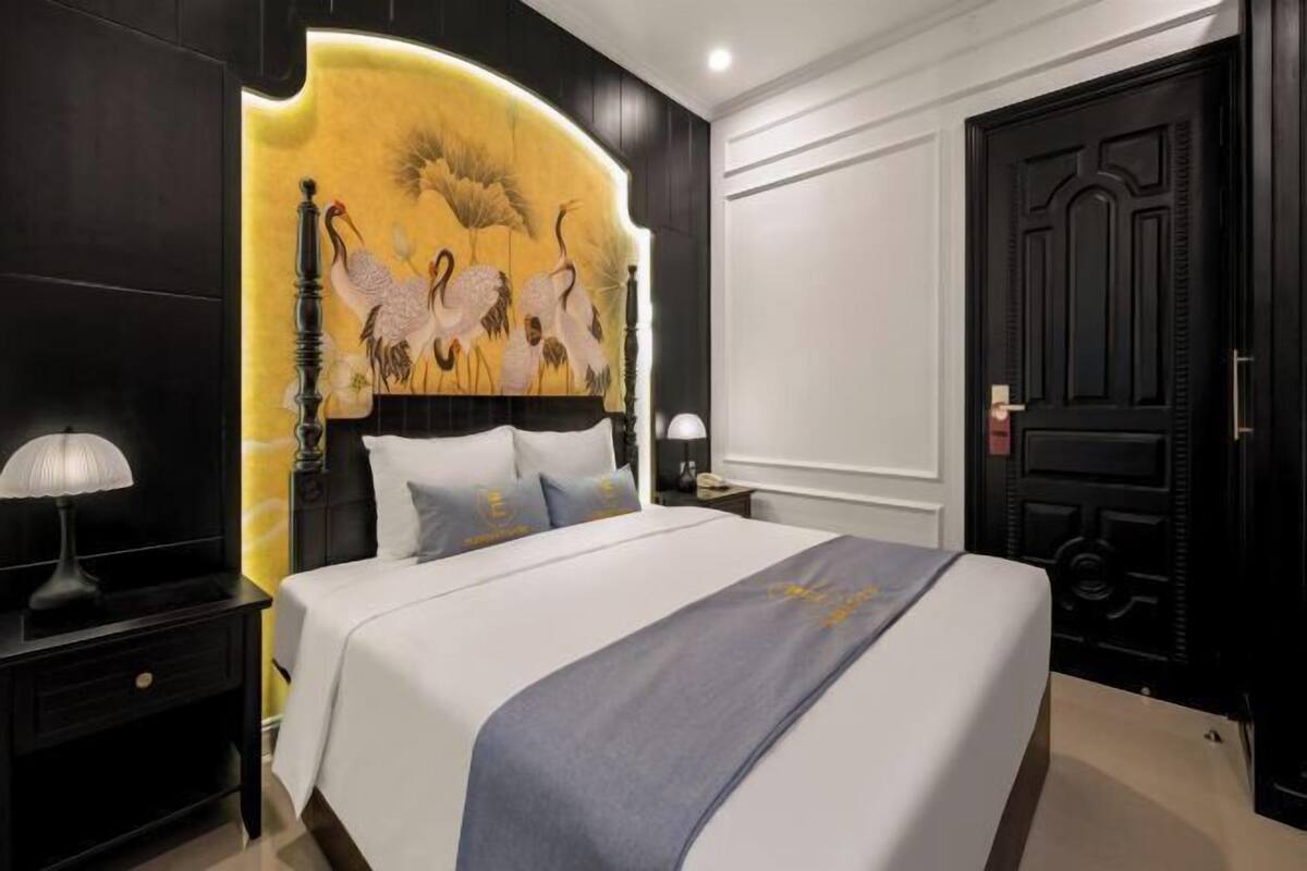 A hotel room with a large bed, a headboard with a painting, a nightstand with a lamp, and a door.