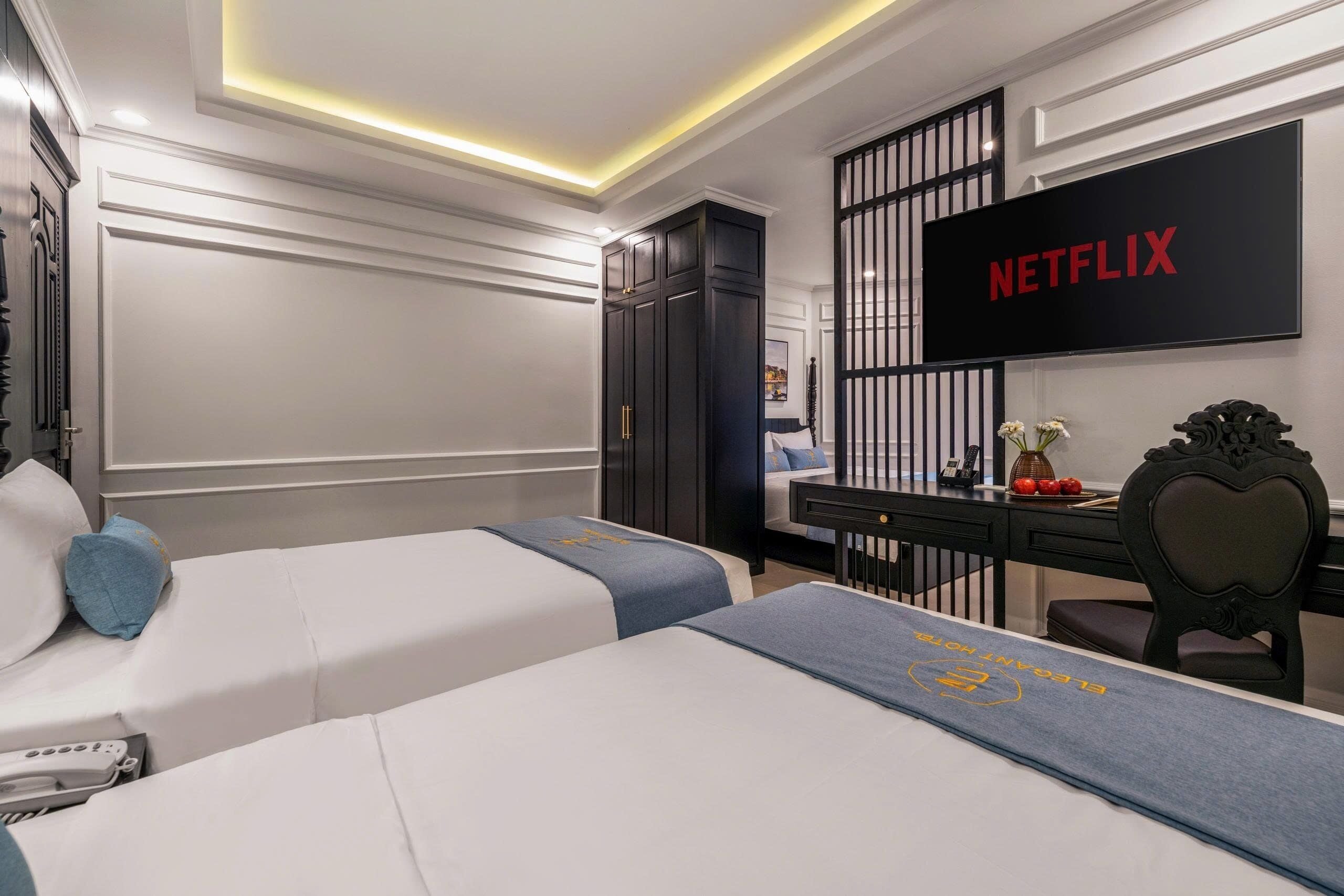 Ein Hotelzimmer mit zwei Betten, einem Schreibtisch, einem Stuhl und einem Fernseher, der 'NETFLIX' anzeigt, sowie einer dekorativen Wandverkleidung.