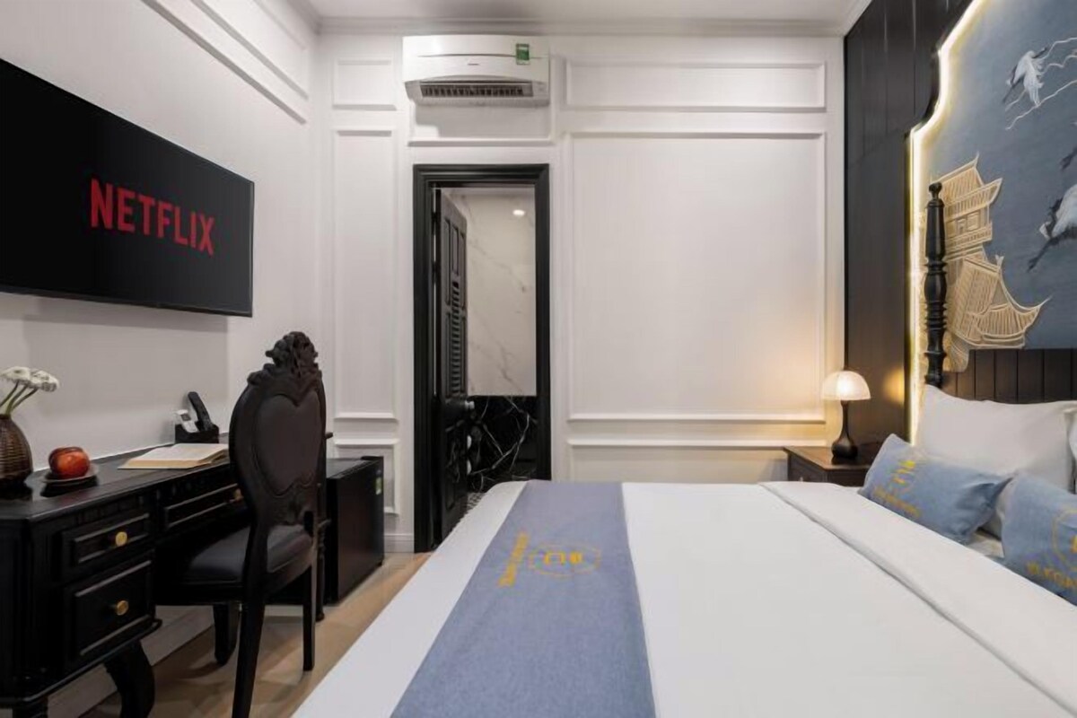 A hotel room with a bed, a desk, a chair, a TV screen, and a wall with a decorative painting.