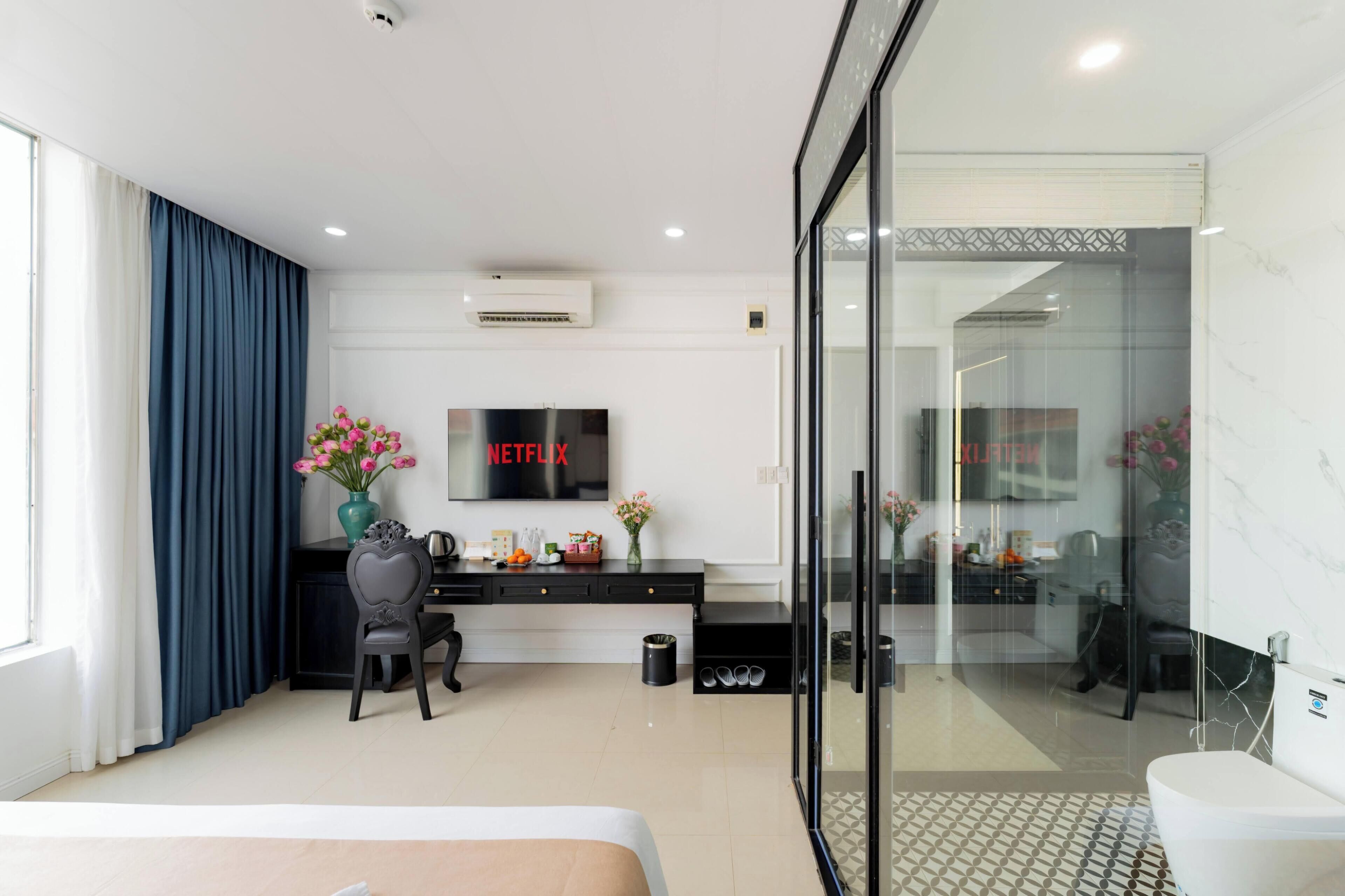 A modern hotel room with a bed, a desk with a chair, a TV mounted on the wall, and a glass shower enclosure.