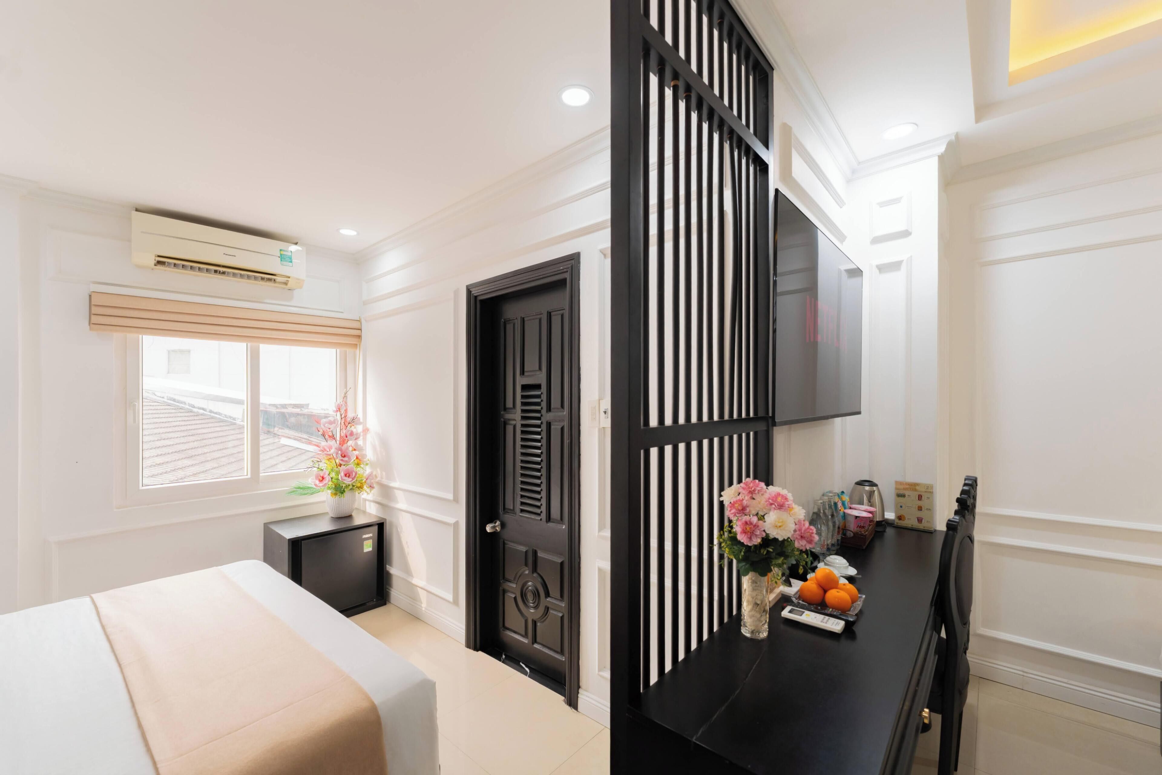 A hotel room with a bed, a desk with a vase of flowers, a black door, and a TV mounted on the wall.