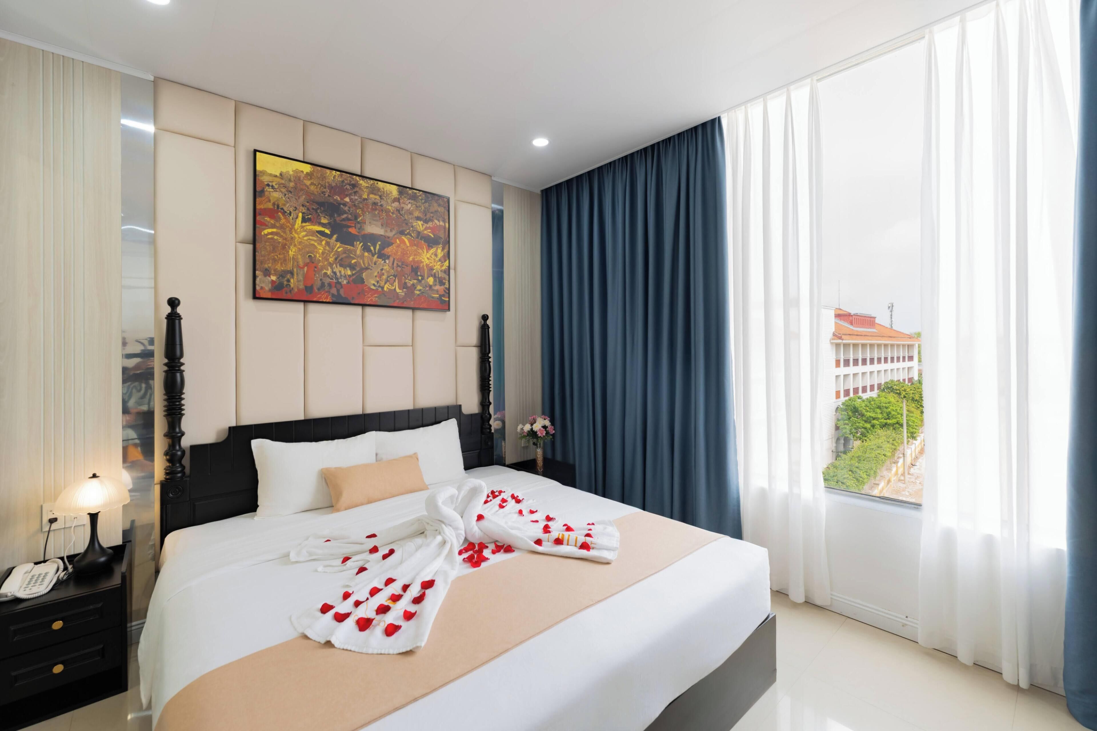 Habitación de hotel con una cama grande, una mesita de noche con lámpara, un cuadro en la pared y vistas al exterior a través de la ventana.