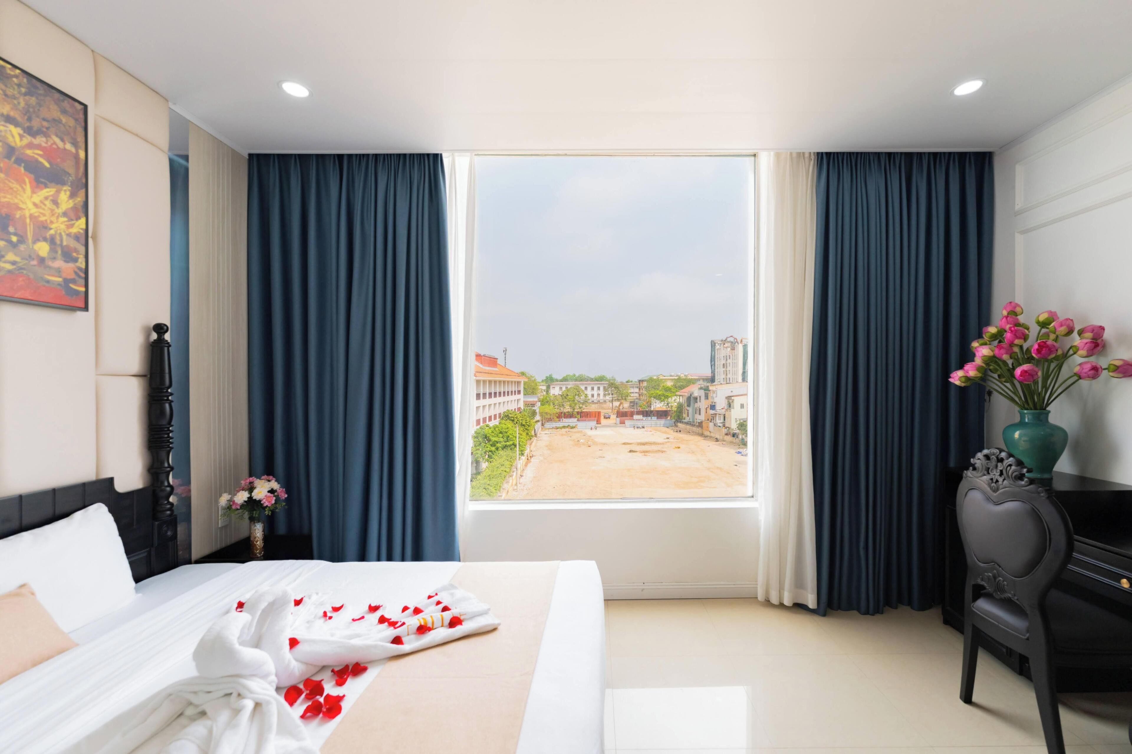 Habitación de hotel con cama, escritorio, silla, un jarrón con flores y vistas a edificios a través de un ventanal grande.