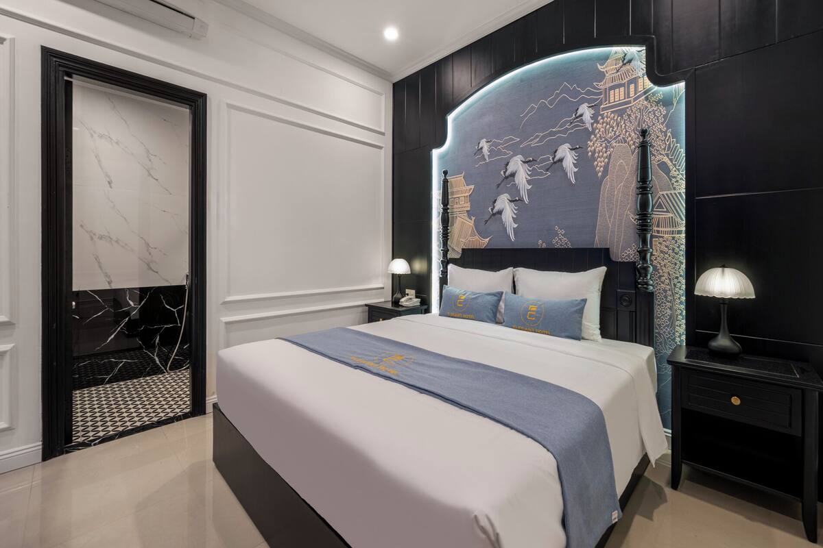 A modern hotel room with a large bed, a headboard with a decorative mural, a nightstand with a lamp, and a door with a marble panel.