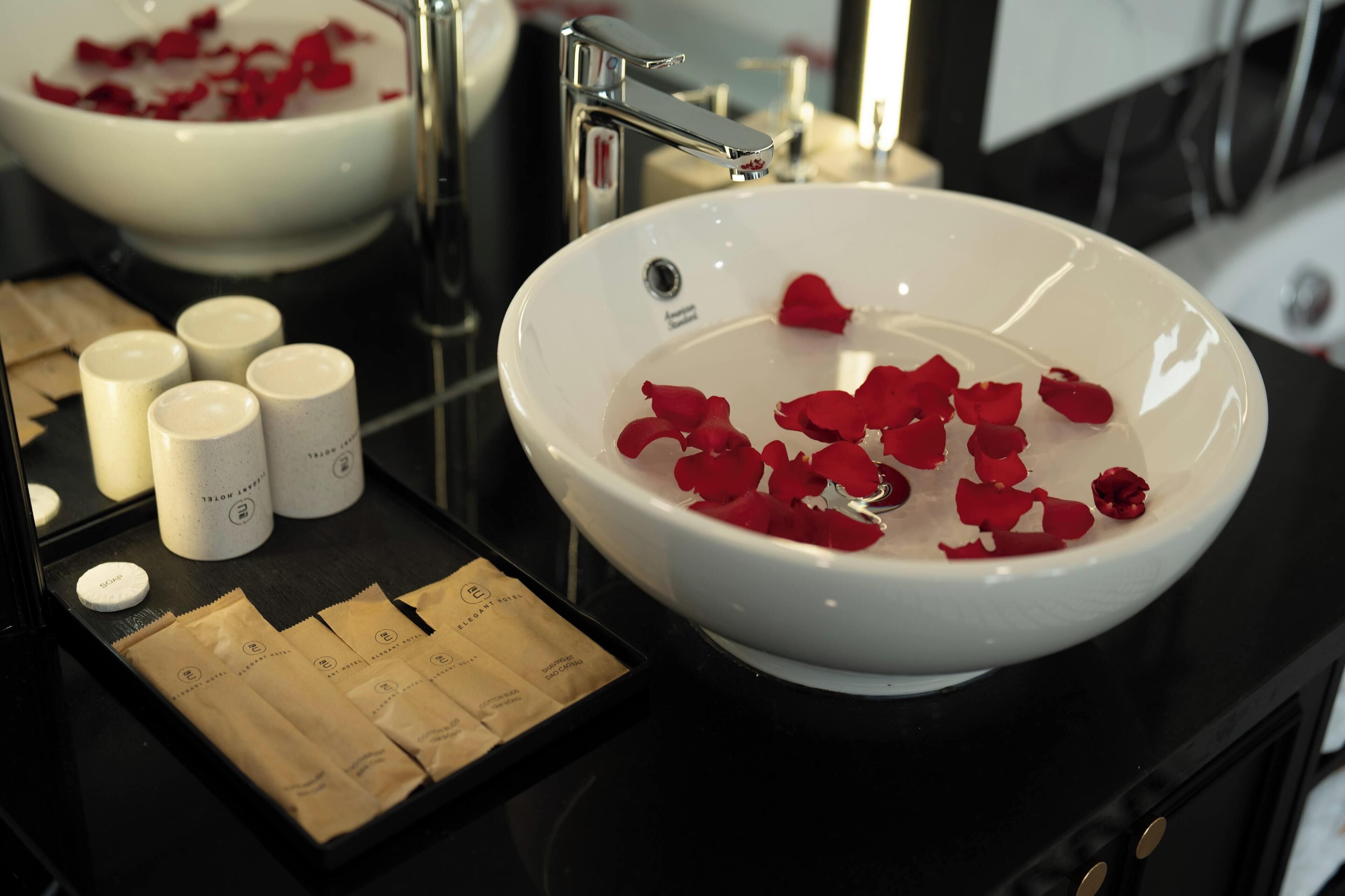 A bathroom with a white sink, rose petals in the water, and toiletries on the counter.