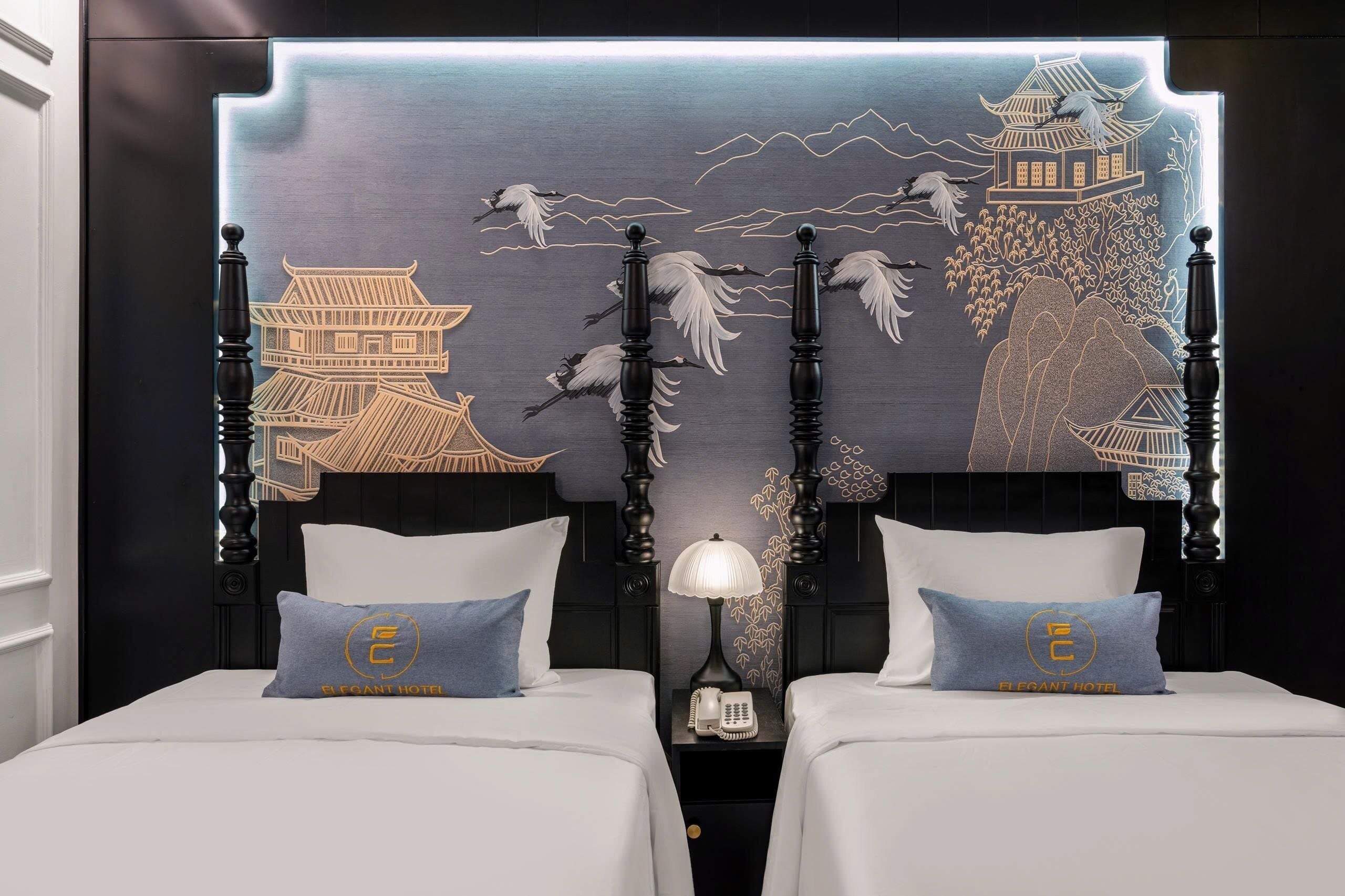 Habitación de hotel con dos camas, una mesita de noche con lámpara y una pared con un mural asiático tradicional.