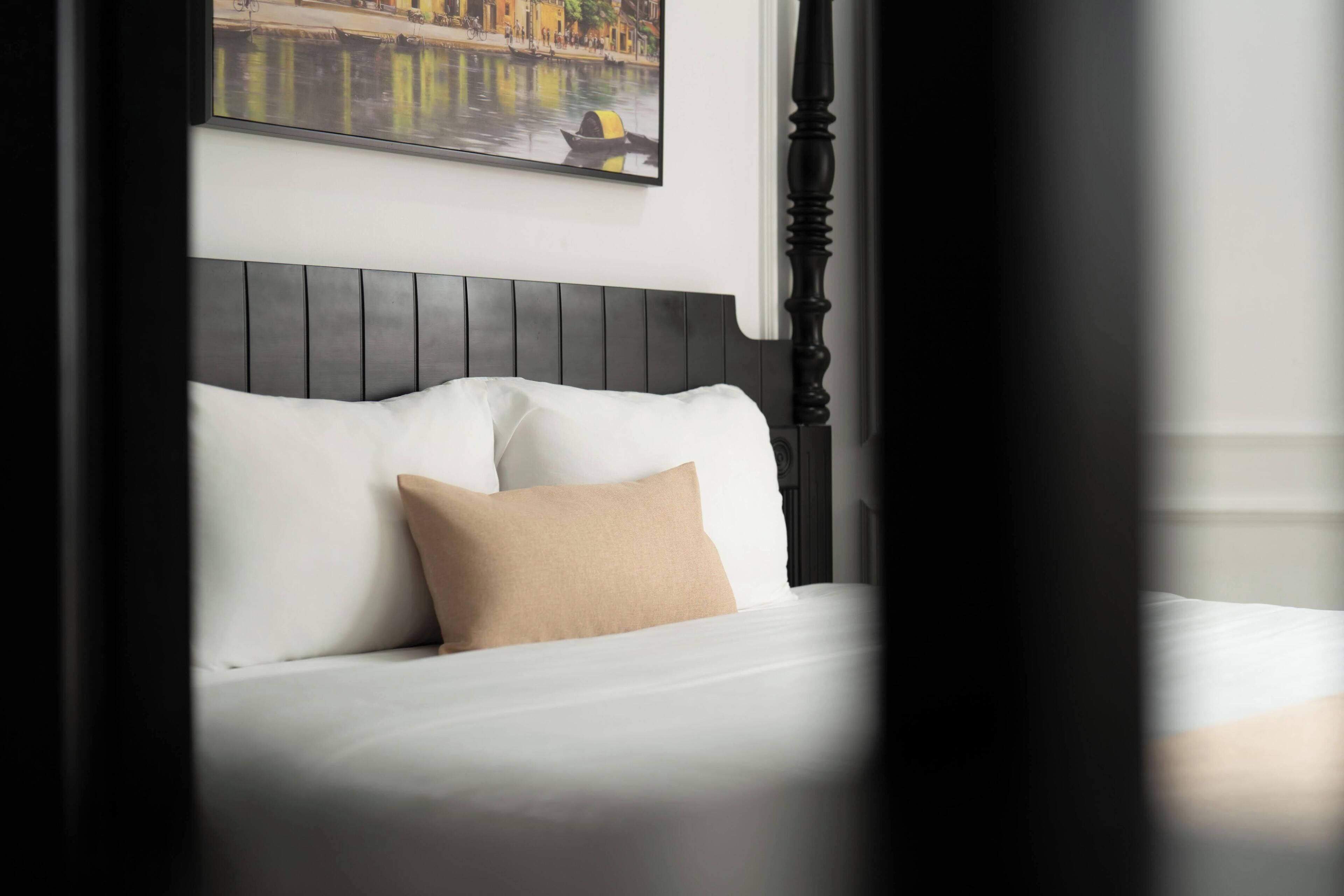 A bed with white bedding and a single beige pillow, a dark wooden headboard, and a painting of a canal scene above it.