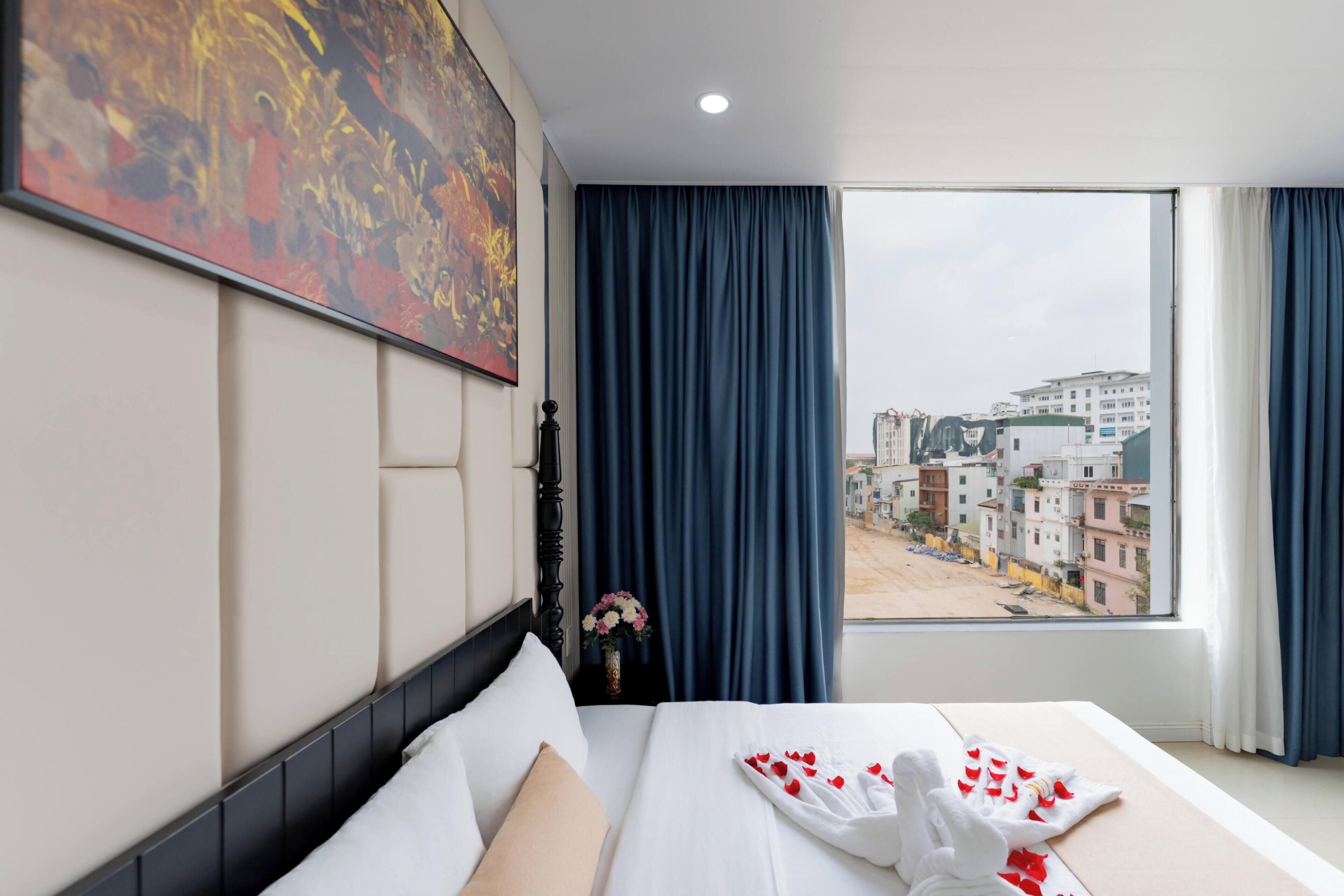 Quarto de hotel com cama, um quadro, janela com vista para edifícios e um vaso com flores.