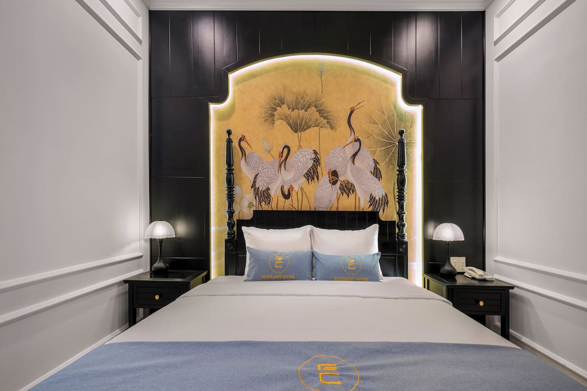 A hotel room with a large bed, a decorative headboard with a bird mural, two bedside tables with lamps, and a blue bedspread with the hotel logo.