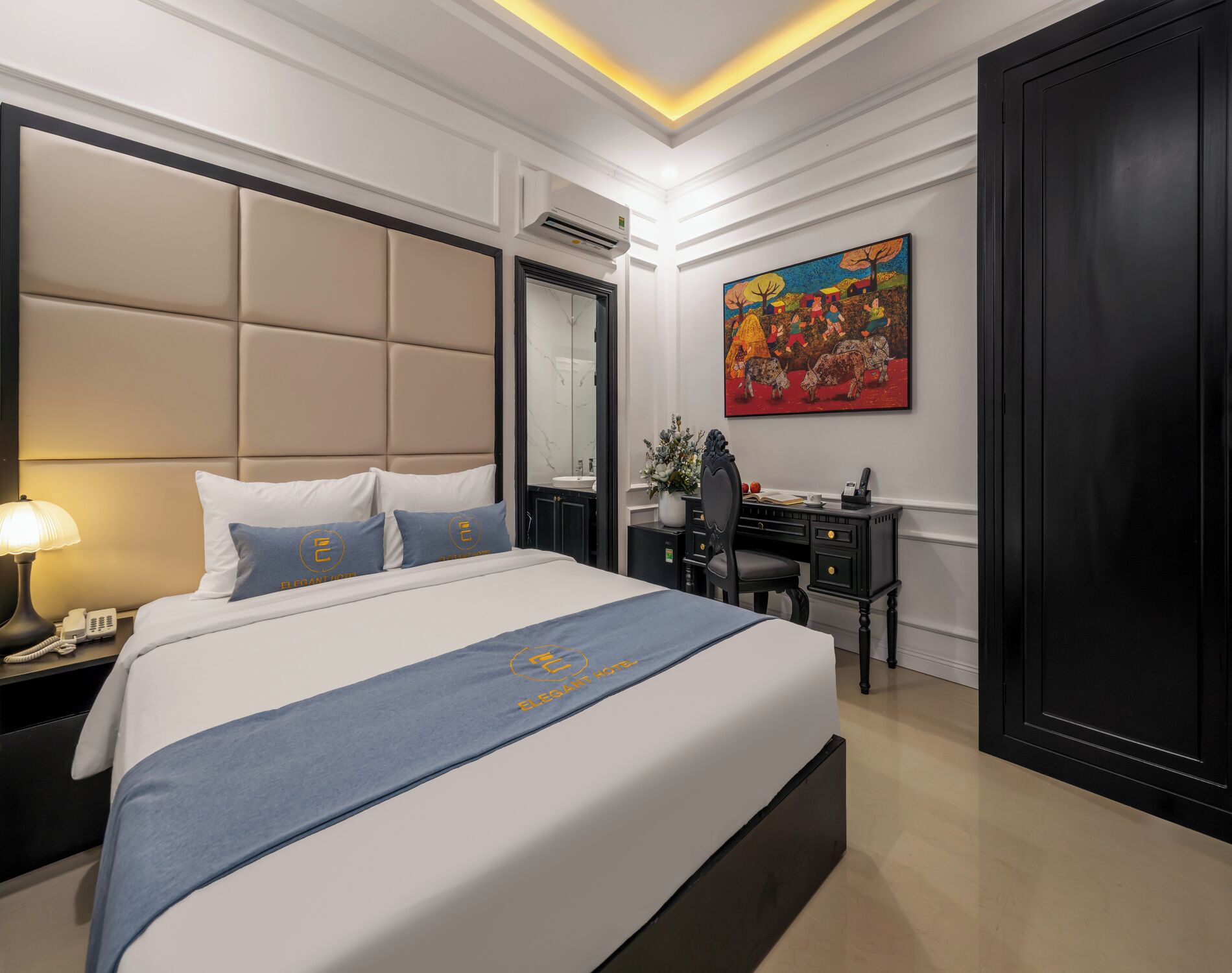 A hotel room with a large bed, bedside table, desk, and a painting on the wall.