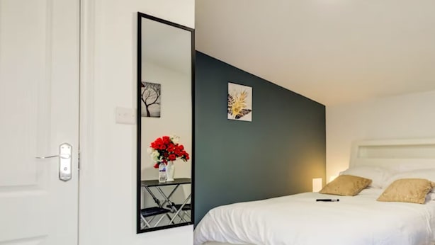 A bedroom with a white bed, a bedside table with a vase of red flowers, a large mirror, and a dark accent wall.