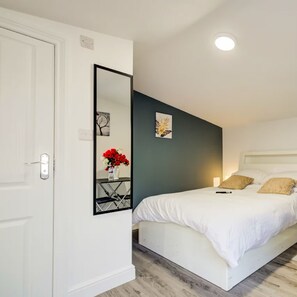 Een slaapkamer met een wit bed, een nachtkastje met een vaas rode bloemen, een grote spiegel en een donkere accentmuur.