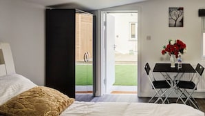 A bedroom with a bed, a desk with chairs, a vase with flowers, and a view of a garden.