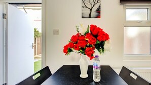A dining area with a table, chairs, a vase with red flowers, a water bottle, and a painting on the wall.