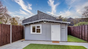 A small, single-story shed with a white door and a window, surrounded by a wooden fence and a well-maintained lawn.
