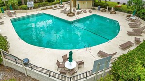 Una piscina con forma de corazón, con sillas de descanso y sombrillas, rodeada de follaje y un pequeño edificio.