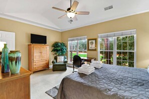 A bedroom with a bed, a ceiling fan, a TV, a wooden cabinet, a chair, a plant, and large windows. - E2 Golfmaster Relaxing Golf Escape with Beach Acce (Hilton Head Island)