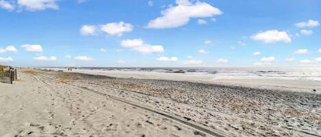 Una playa con un sendero arenoso que lleva al agua, una valla de madera a la izquierda y un cielo despejado con nubes.