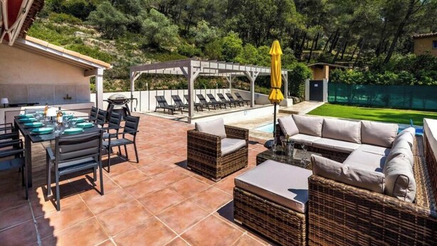 Eine Außenpatio mit Esstisch, Stühlen und einer Couch. Eine Pergola mit einem gelben Sonnenschirm steht dort, im Hintergrund ist ein Pool zu sehen.