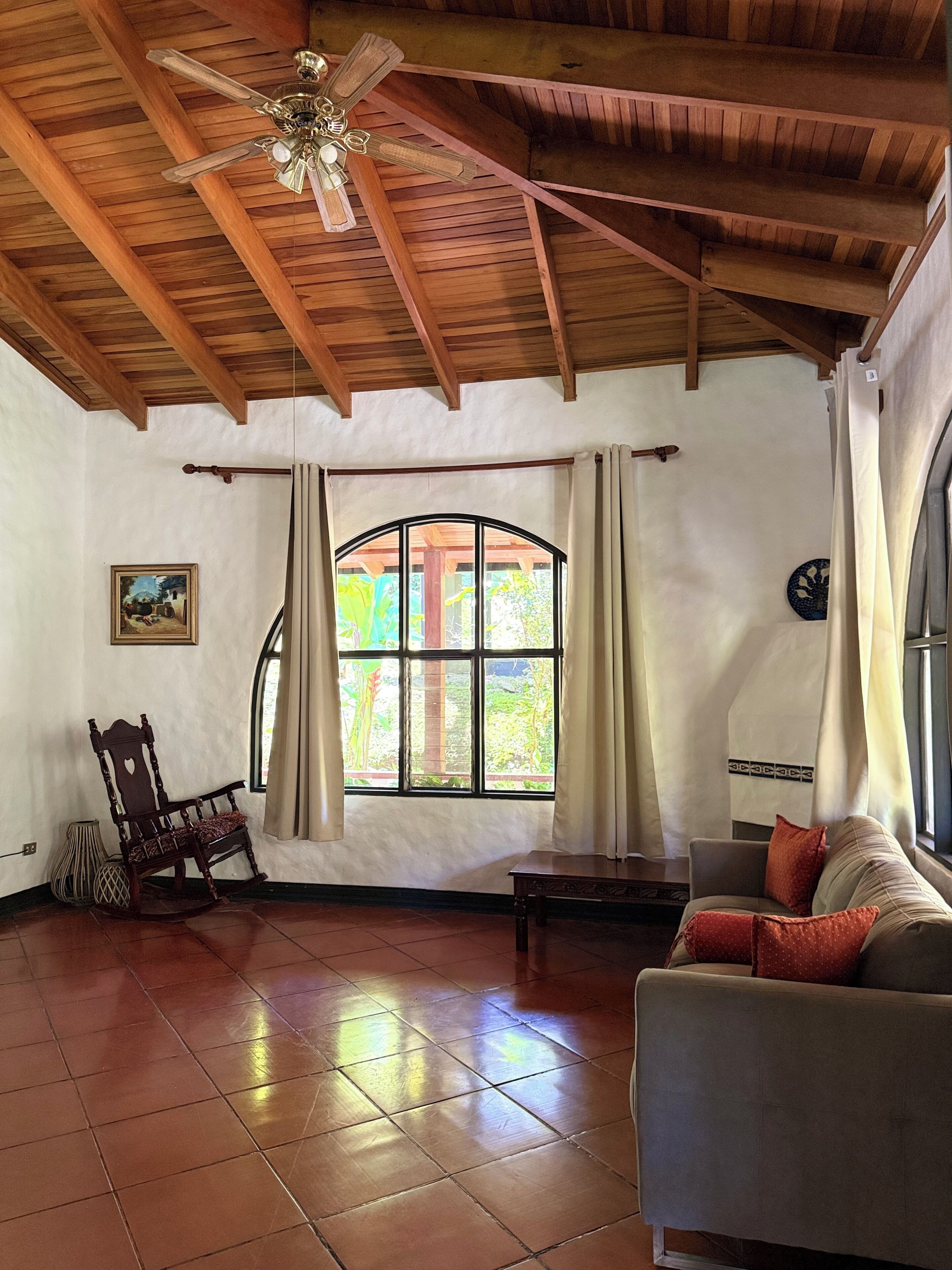 Una habitación con techo de madera, un ventanal con cortinas, un sillón mecedor, un sofá y una mesa.