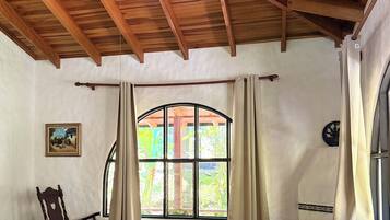 Una habitación con techo de madera, un ventanal con cortinas, un sillón mecedor, un sofá y una mesa.
