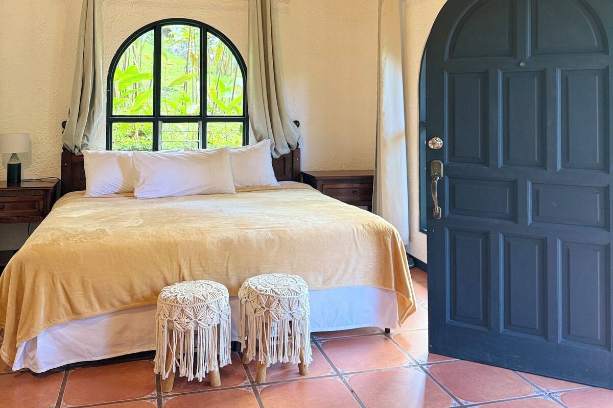 A bedroom with a large bed, two macramé poufs, a wooden ceiling, a large window, and a dark blue door.