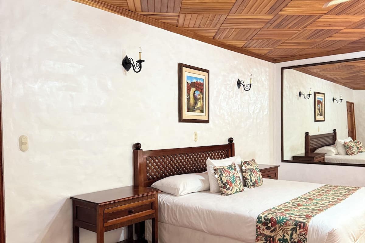 A bedroom with a wooden ceiling, a bed with white bedding, a nightstand, a wall-mounted lamp, and a framed picture.
