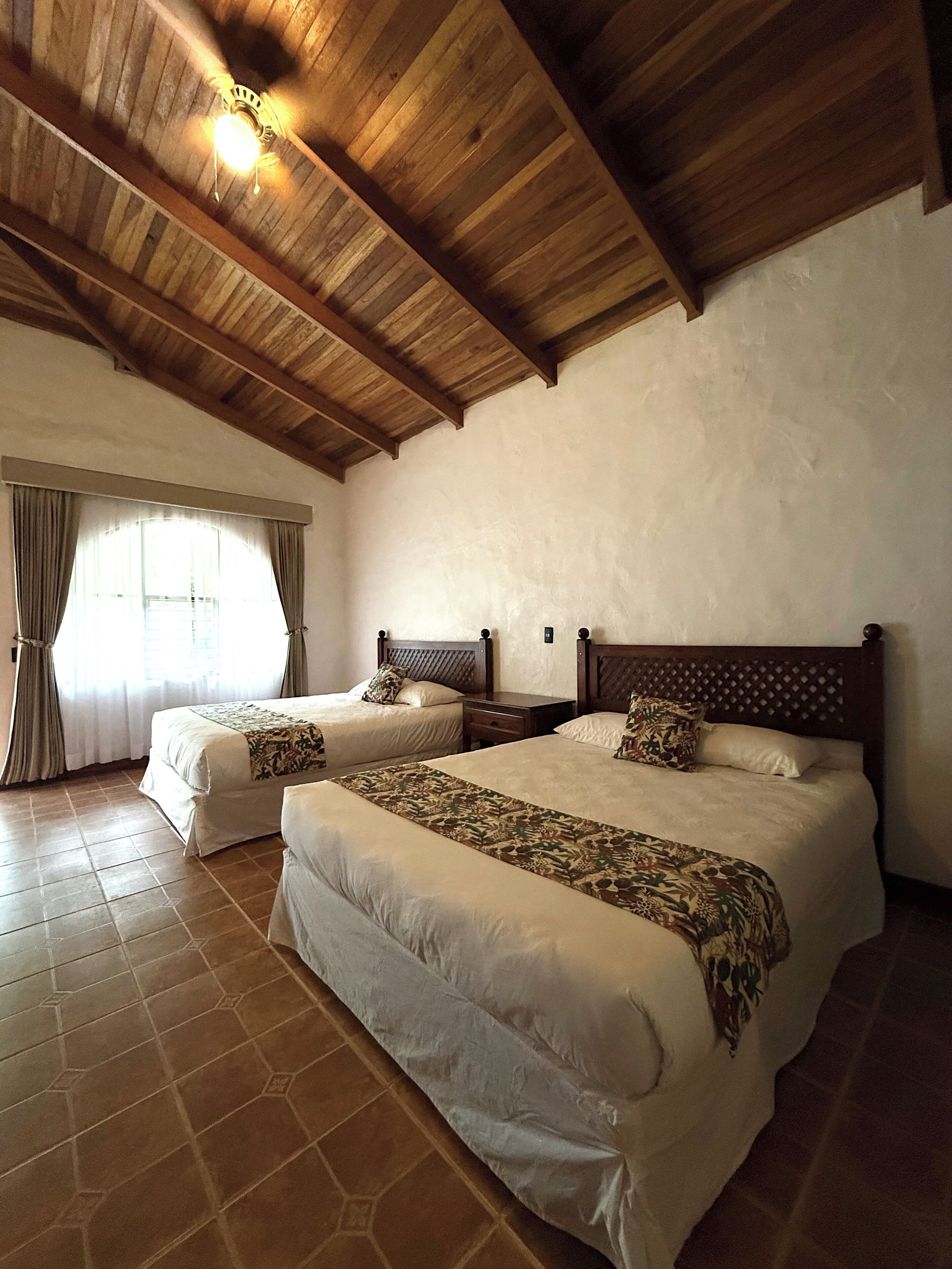 A hotel room with two beds, a wooden ceiling, and a large window with curtains.