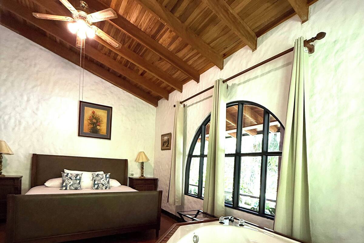 A bedroom with a large bed, a wooden ceiling, a ceiling fan, a window with curtains, and a bathtub.