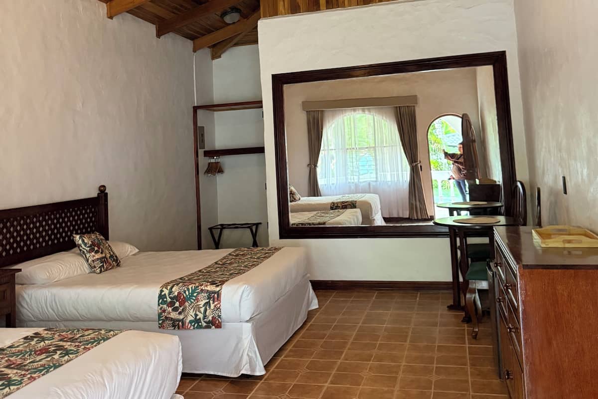 A room with two beds, a wooden ceiling, a large mirror, and a view through a window.
