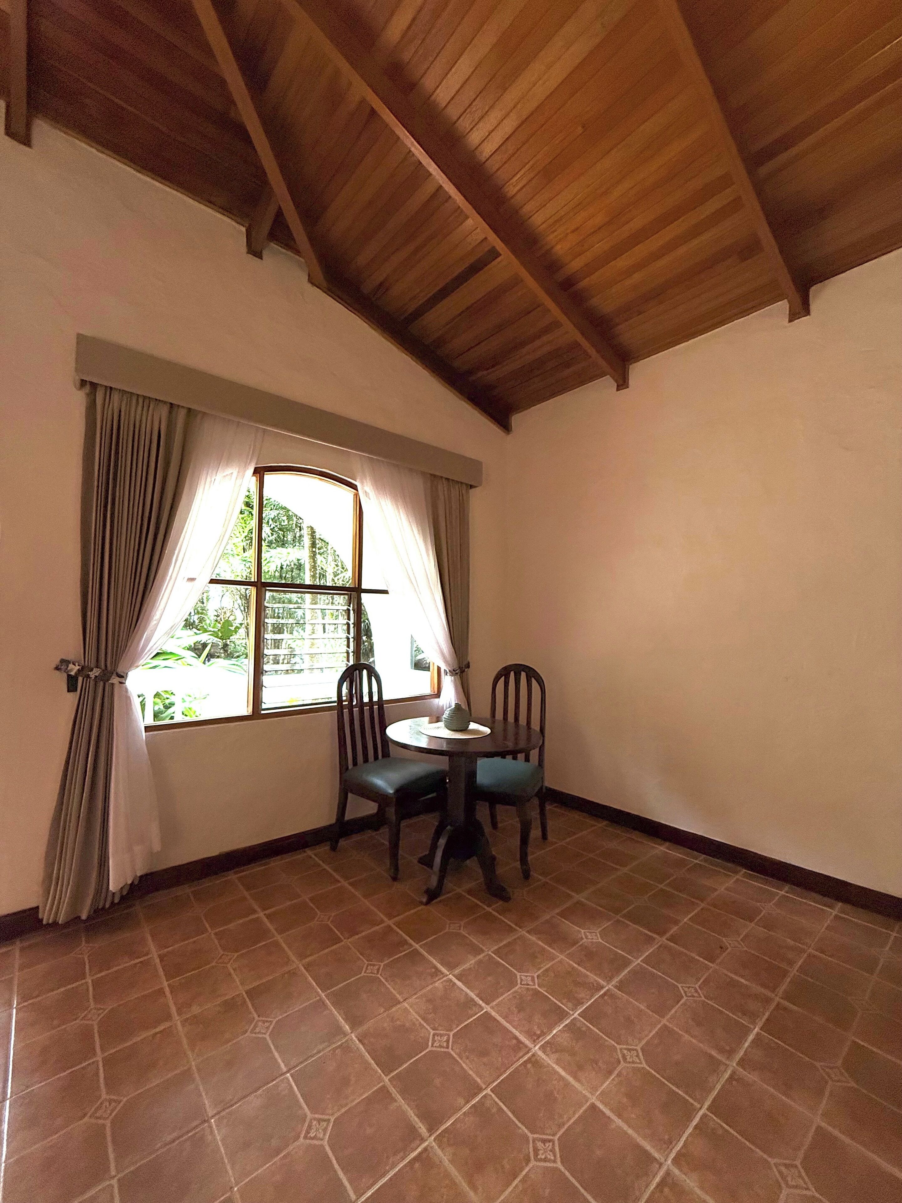 Una habitación con suelo de baldosas, una ventana con cortinas, una mesa redonda con sillas y techo de madera.