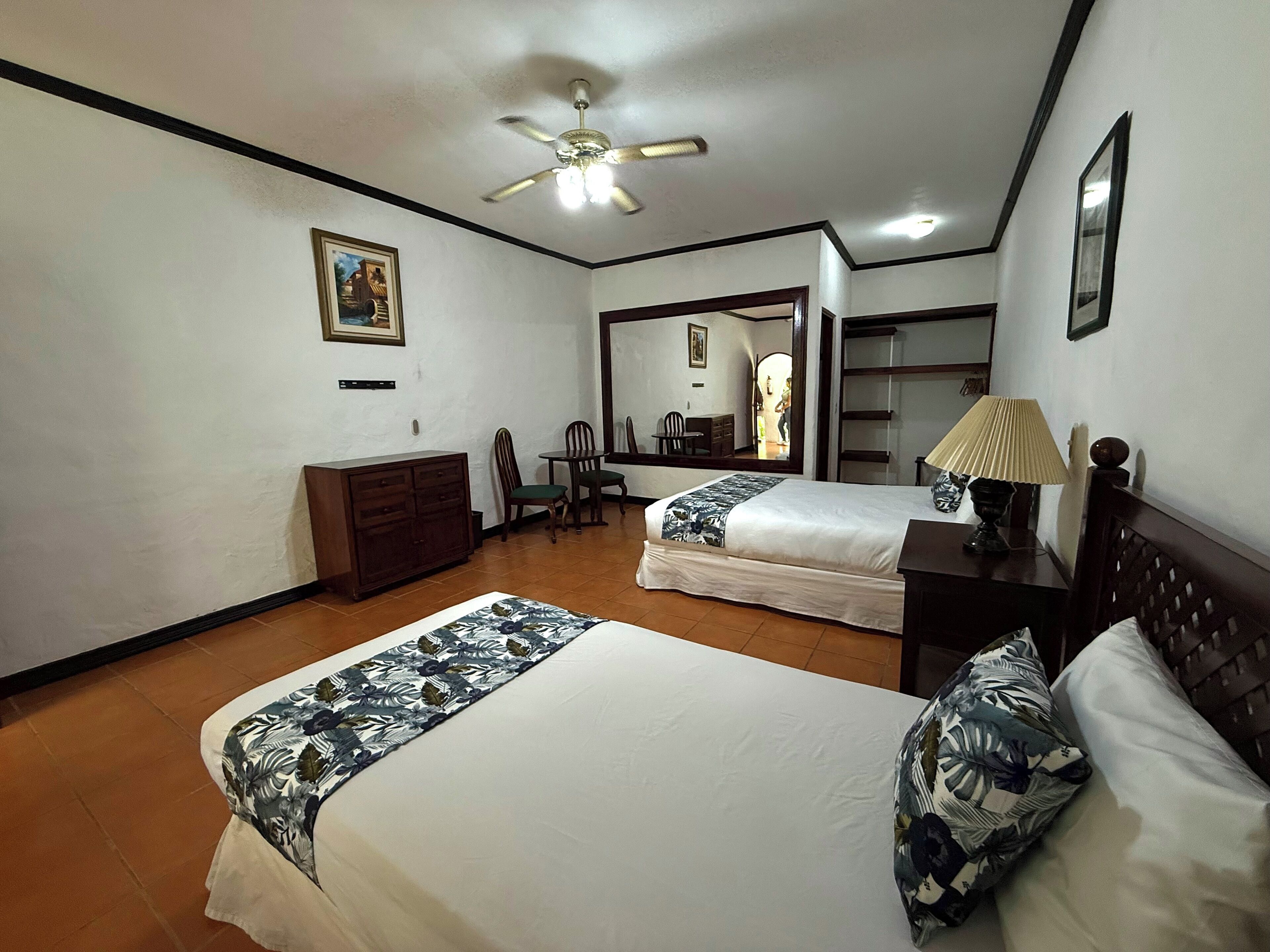 A hotel room with two beds, a desk, a chair, a dresser, a ceiling fan, and a mirror.