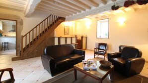 A spacious living room with a black leather sofa, a wooden coffee table, and two leather armchairs. A wooden staircase leads to a kitchen area.