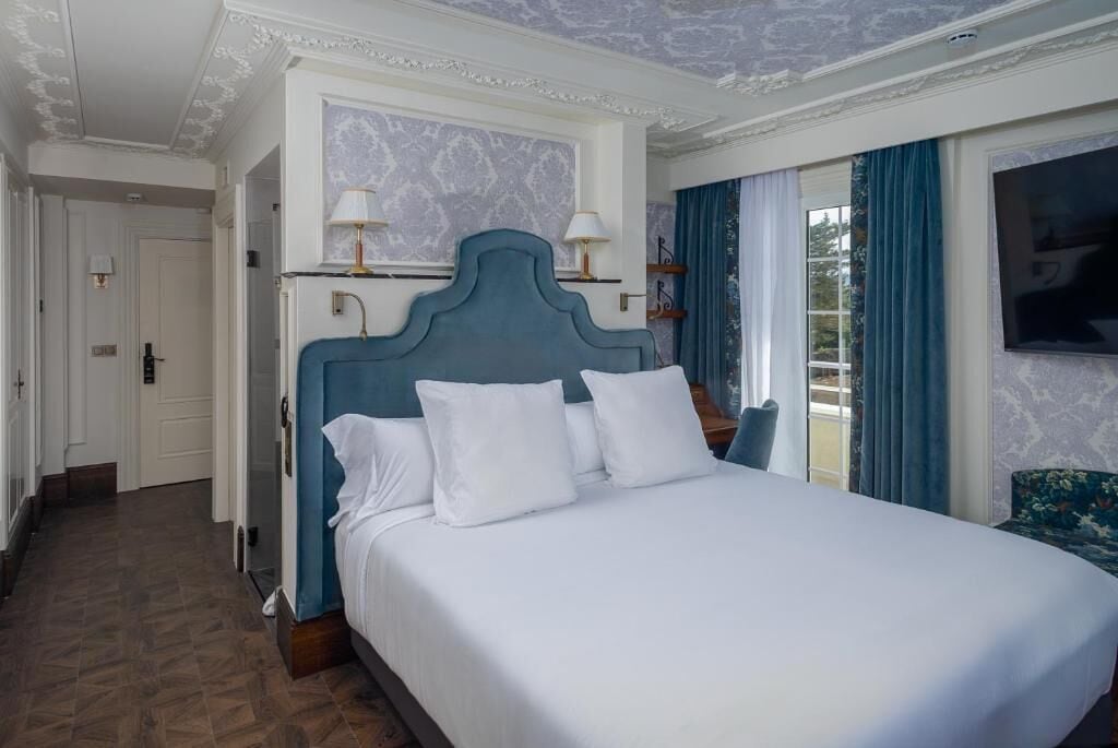 Habitación de hotel con una cama grande, cabecera, dos lámparas de noche, televisor y vistas al exterior.