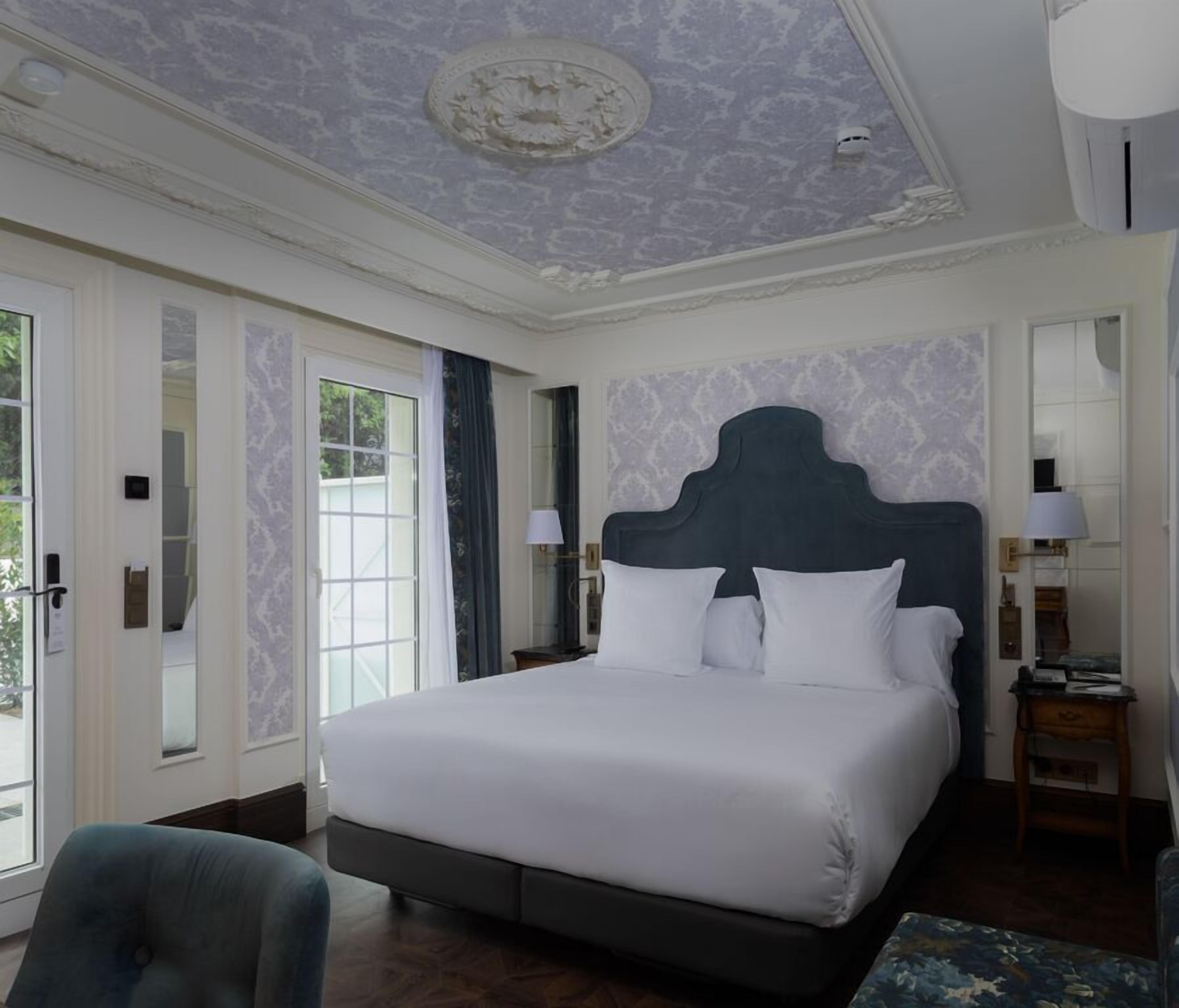 Una cama grande con cabecera oscura, una mesita de noche con lámpara y una silla en una habitación con techo estampado y ventanas amplias.