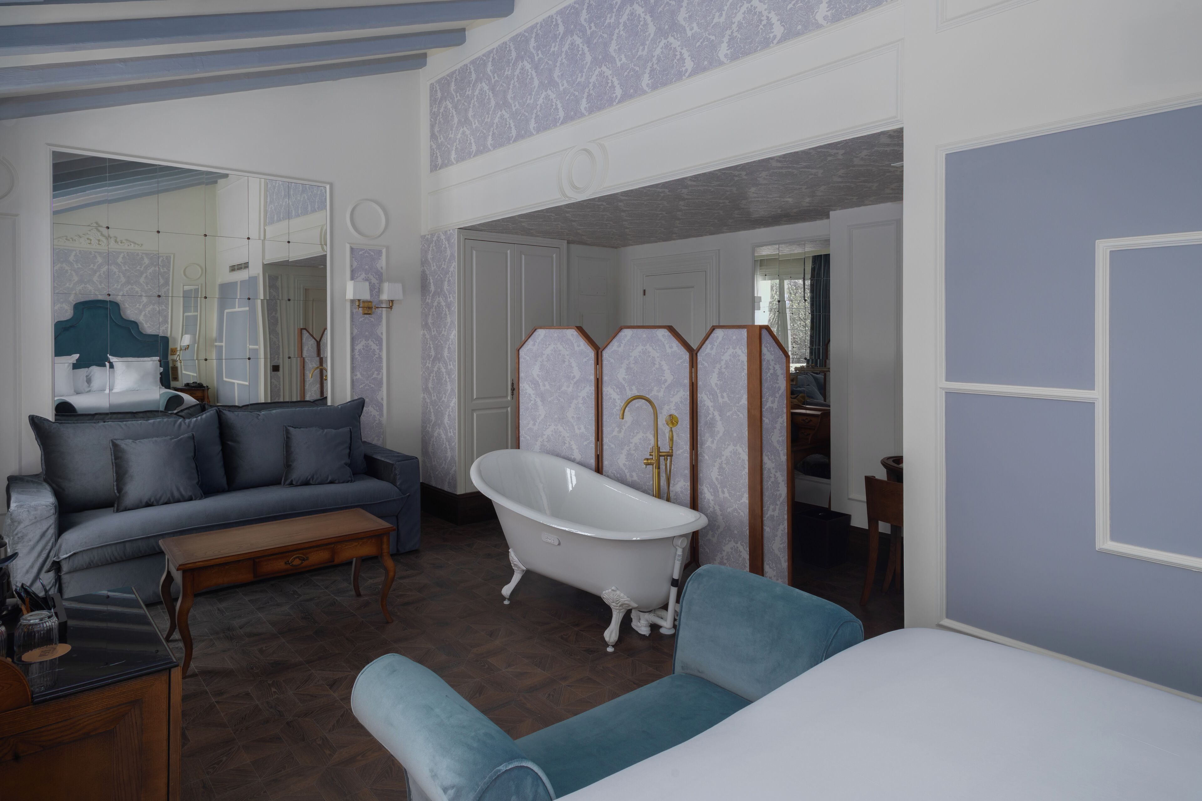 A room with a blue sofa, a wooden coffee table, a white bathtub, and a mirror.