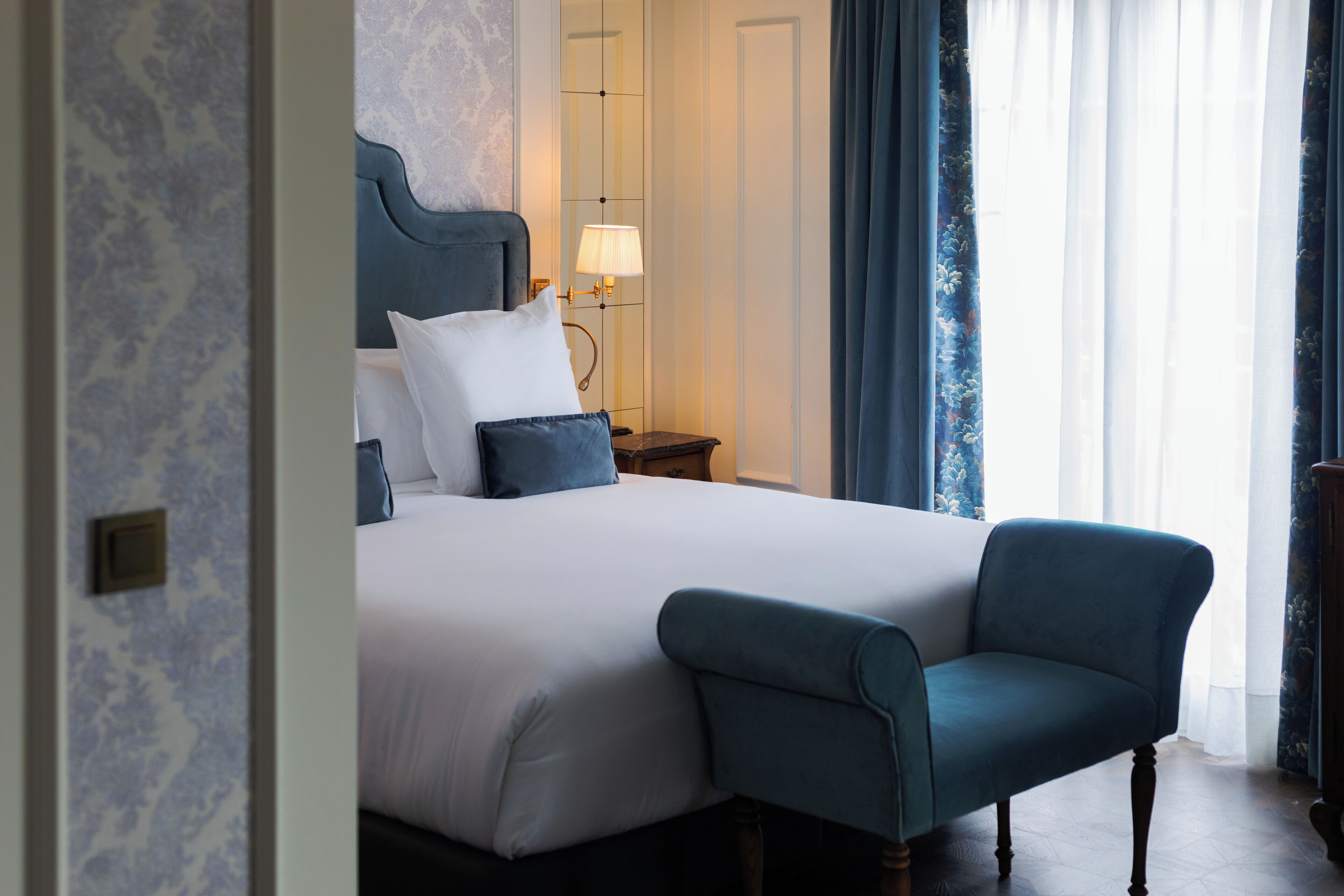 Habitación de hotel con una cama grande, una mesita de noche con lámpara, una silla y vistas al exterior a través de cortinas traslúcidas.