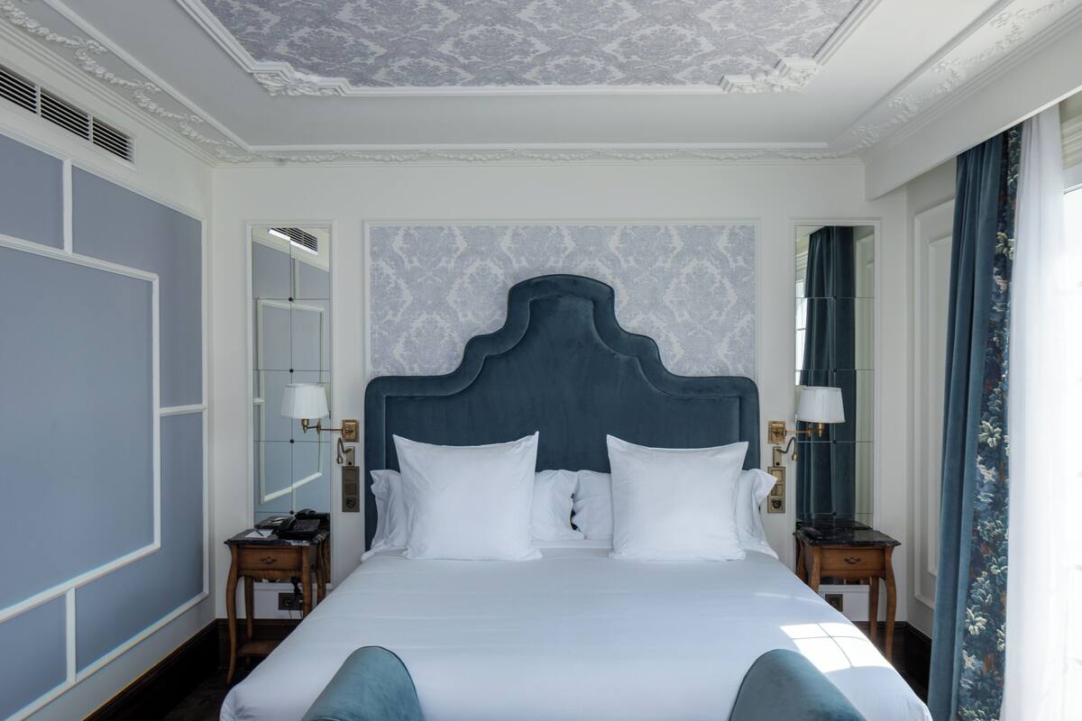 Una cama grande con cabecera oscura, ropa de cama blanca y una silla de color azul. Hay dos mesitas de noche con lámparas, un espejo y un techo con diseño.