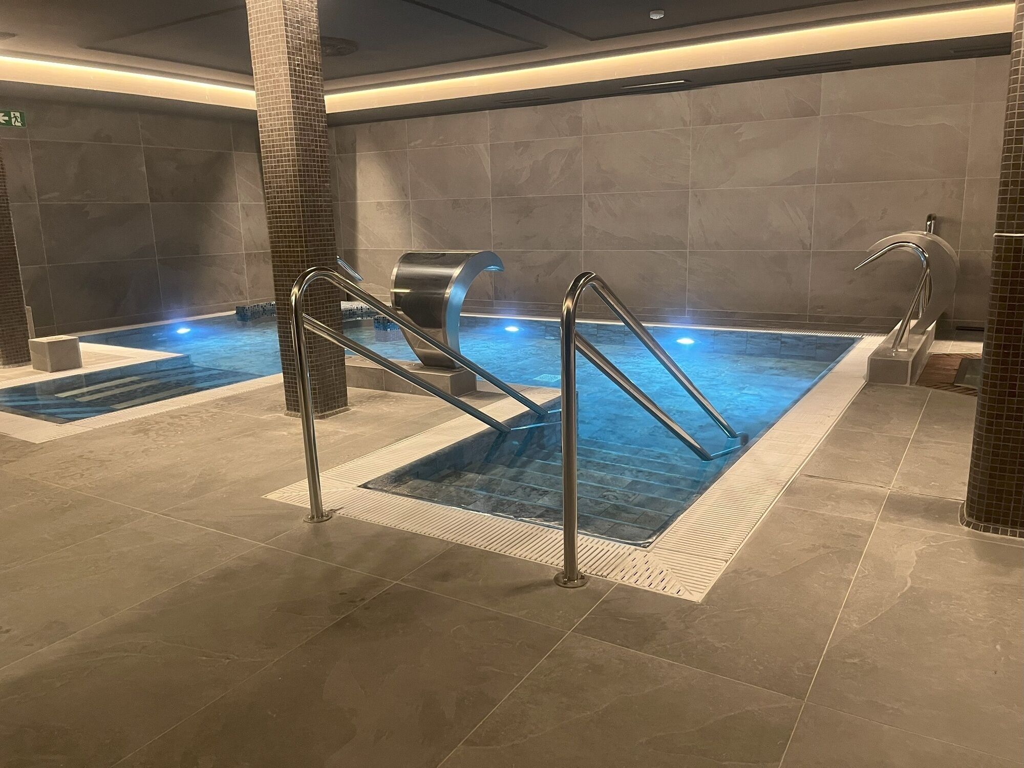 Una piscina interior con agua cristalina, barandales de metal y suelo de baldosas.