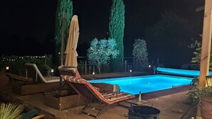 Een zwembad buiten met lounge stoelen en een overdekte parasol, omgeven door hoge bomen en sfeerverlichting.