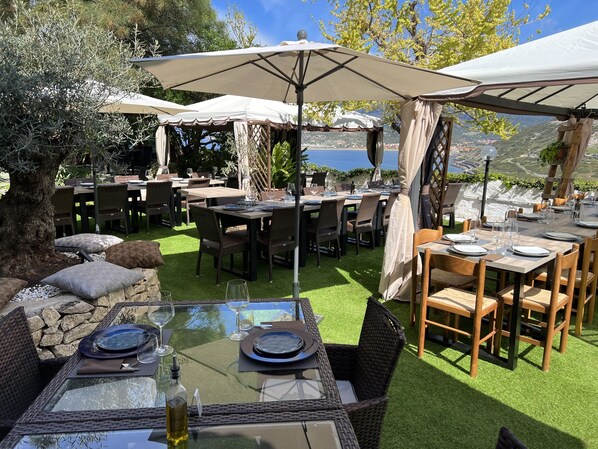 An outdoor dining area with tables and chairs, umbrellas, and a view of the sea.