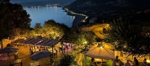 An outdoor dining area with tables and umbrellas, overlooking a coastal landscape with a road and distant city lights.
