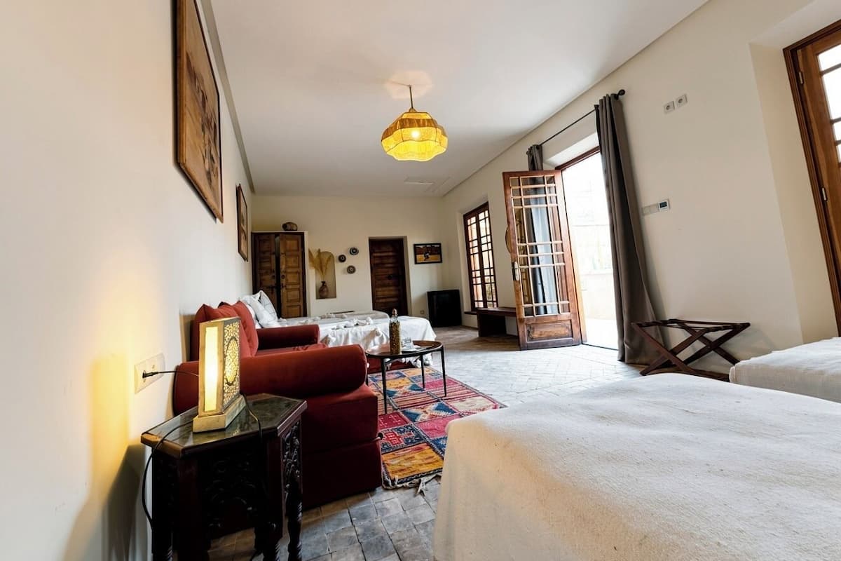 A room with a bed, a sofa, a small table, a lamp, and a window with curtains.