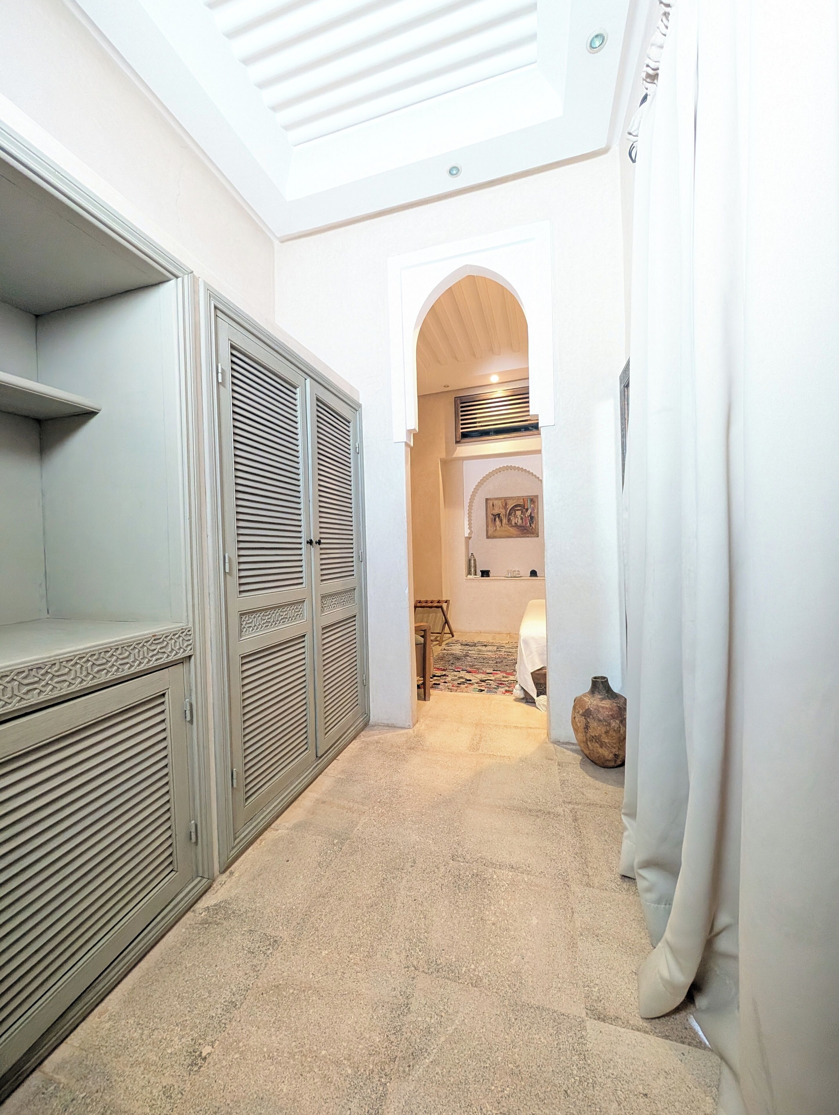 A narrow hallway with a high ceiling, a patterned floor, and a large arched doorway leading to another room.
