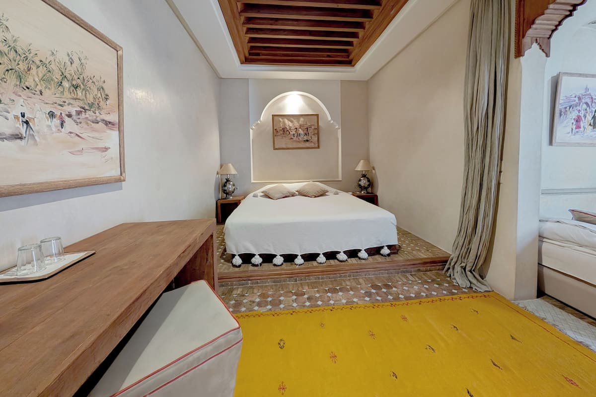 A hotel room with a bed, bedside tables, a wooden bench, a yellow rug, and a painting on the wall.