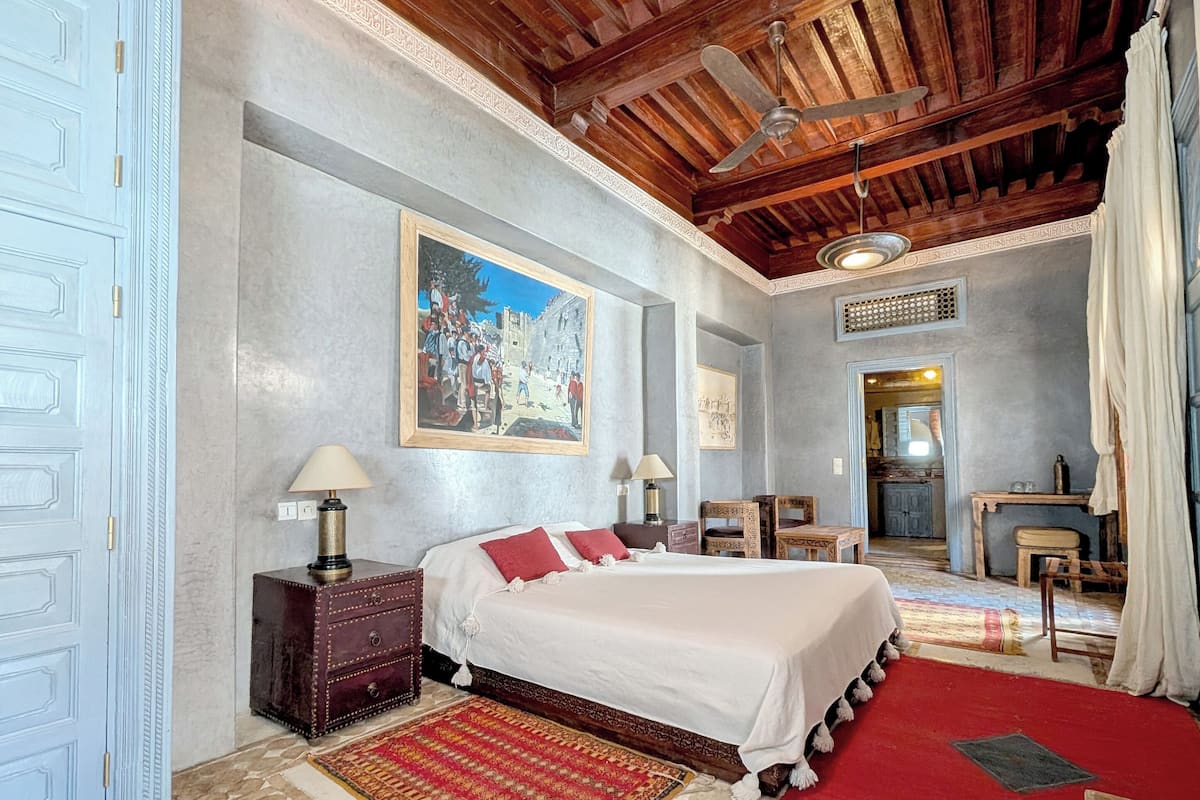 A bedroom with a large bed, a wooden ceiling, a painting on the wall, and a red rug.