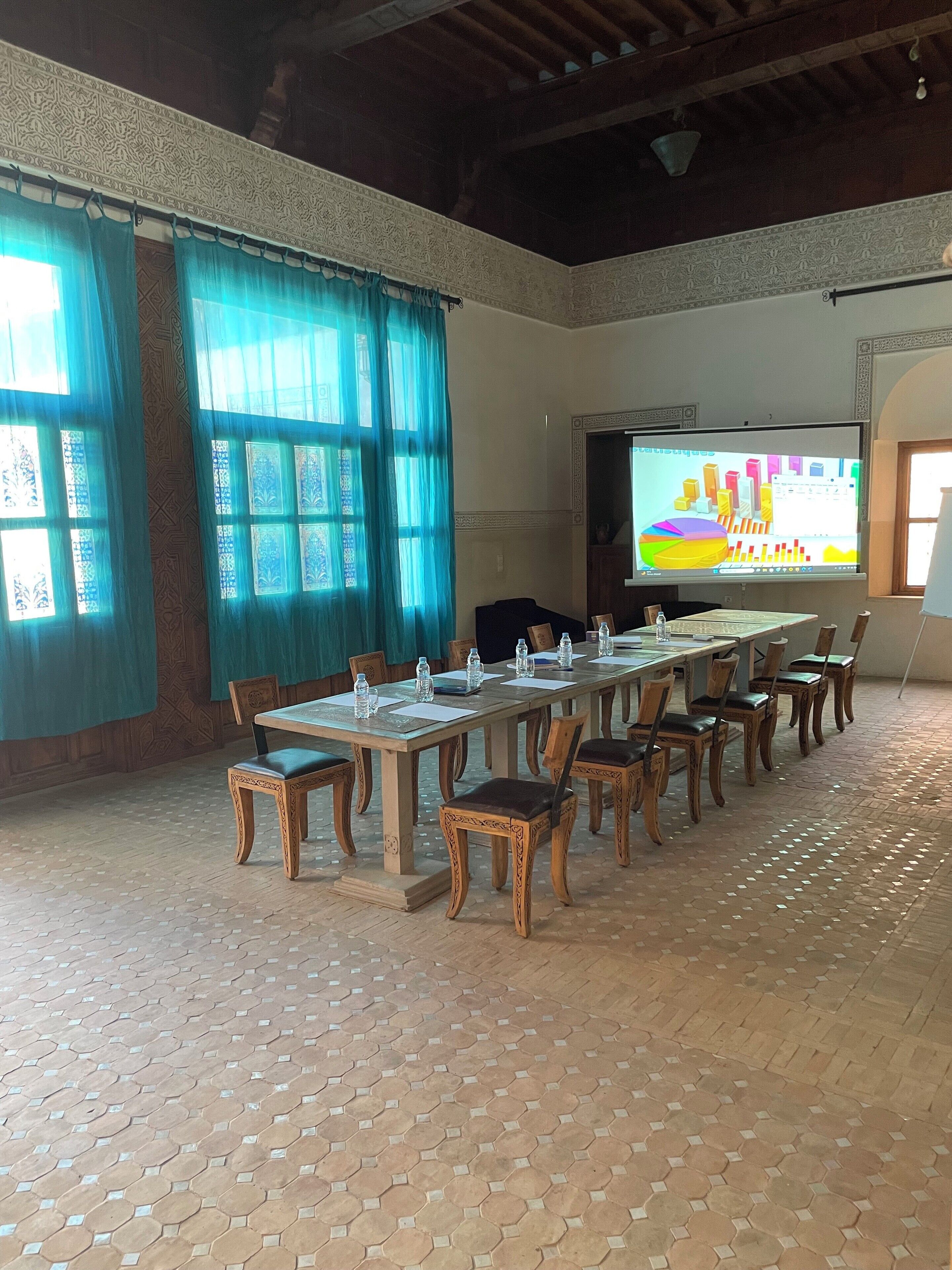 Una sala de conferències amb una taula llarga, cadires, una pantalla projectant gràfics colorits i finestres amb cortines blaves de disseny.