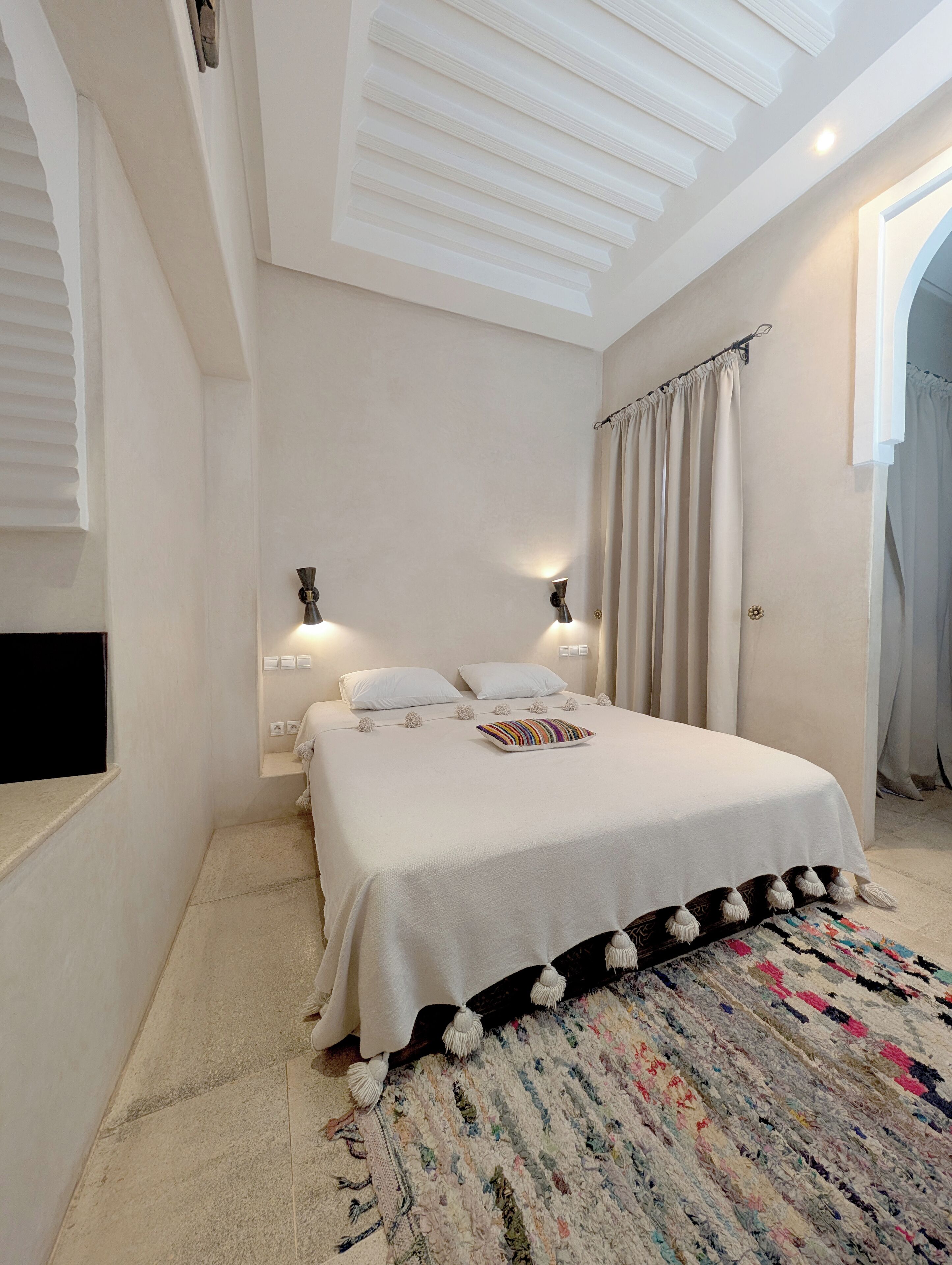 Un dormitorio con cama, dos lámparas de noche, un televisor montado en la pared, un tapete estampado y una ventana con cortinas.