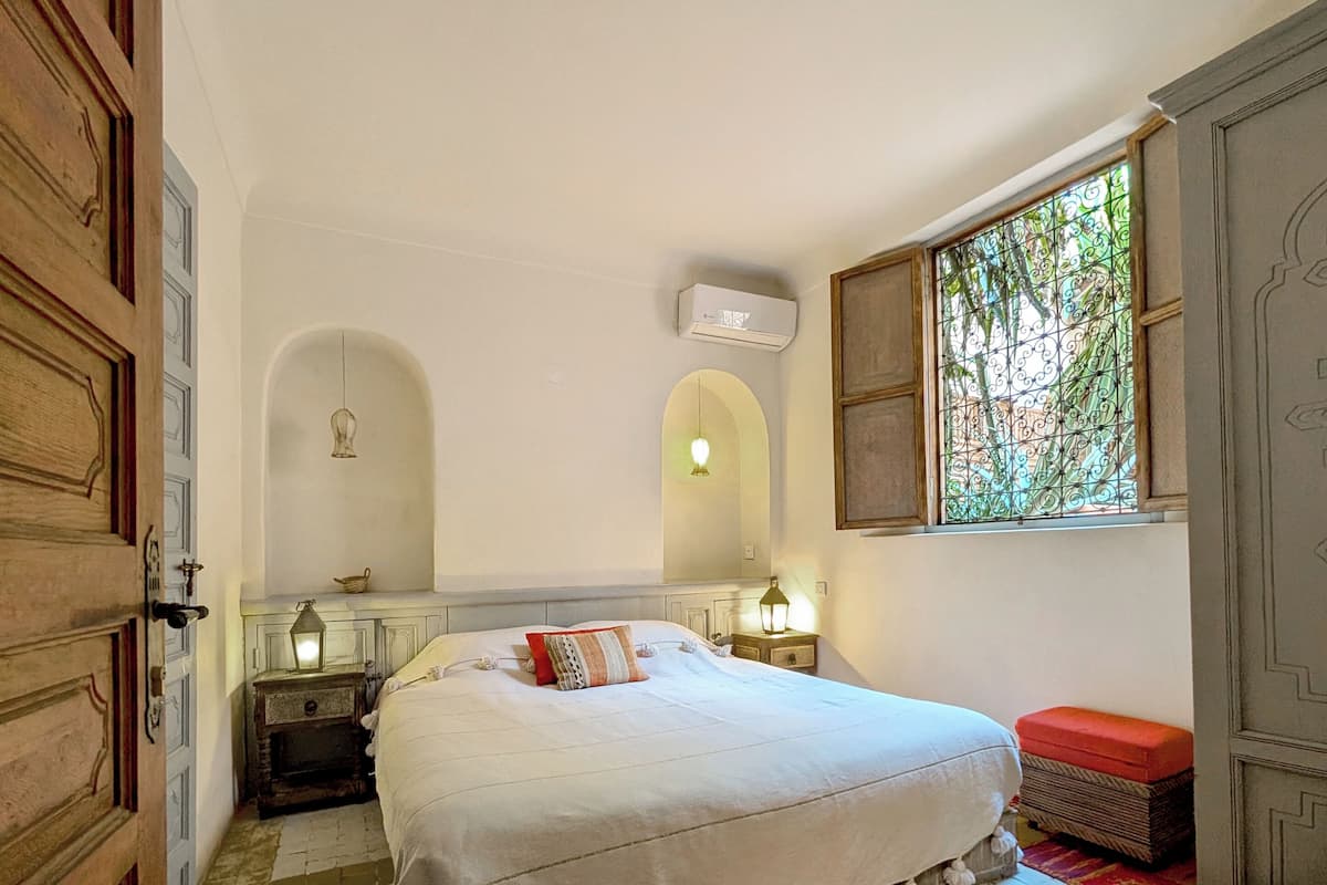 A bedroom with a large bed, a wooden wardrobe, a small table, and a window with decorative glass.