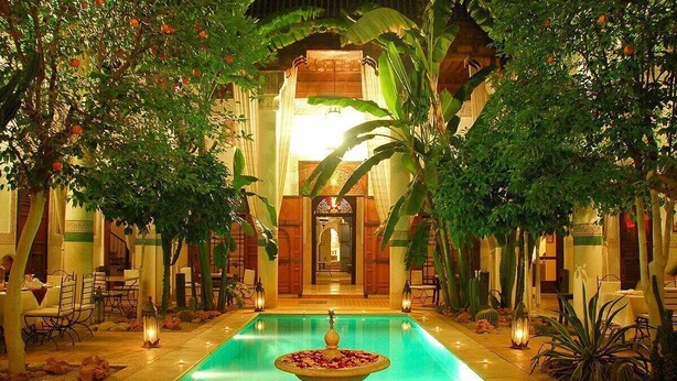 Un espace piscine agrémenté d’une fontaine, entouré d’une végétation luxuriante et bordé d’édifices à l’architecture traditionnelle.